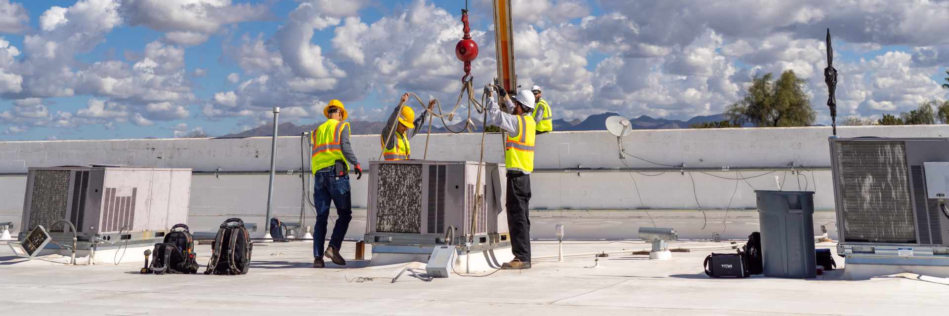 Four workers in hard hats and hi-vis vests on a white commercial roof guiding rigging straps onto a packaged unit under a crane hook