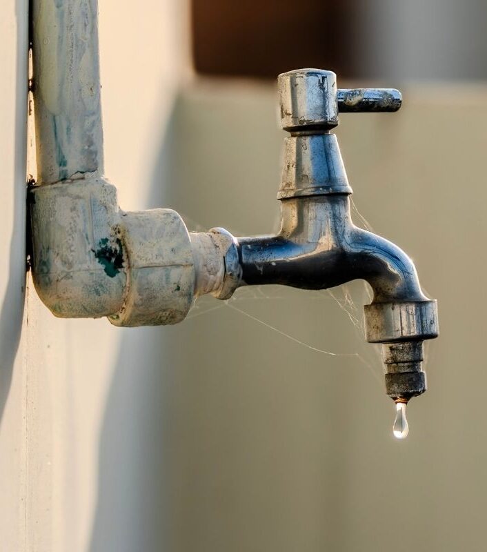 Weathered outdoor spigot with mineral scale on the fitting and a drop of water hanging from the outlet