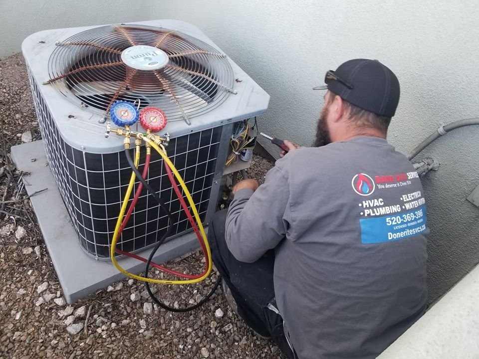 Technician in a Done Rite Services shirt and cap crouched at a residential condensing unit with red and yellow refrigerant gauge hoses connected to the service ports