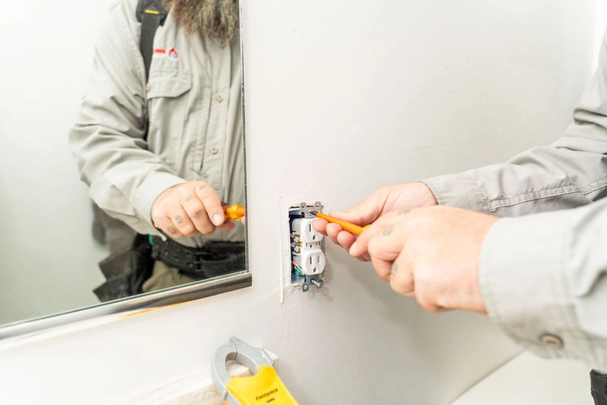 Electrician's hands easing a receptacle out of a wall box beside a bathroom mirror with a clamp meter resting on the counter below