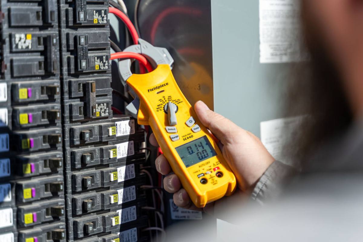 Hand holding a yellow clamp meter against a column of breakers inside an open panel