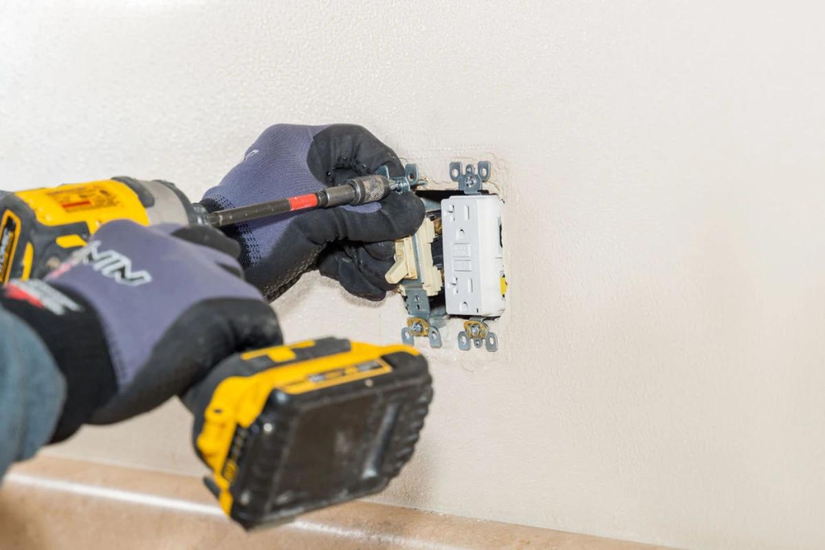 Gloved hands driving a screw into a white GFCI receptacle with a cordless impact driver