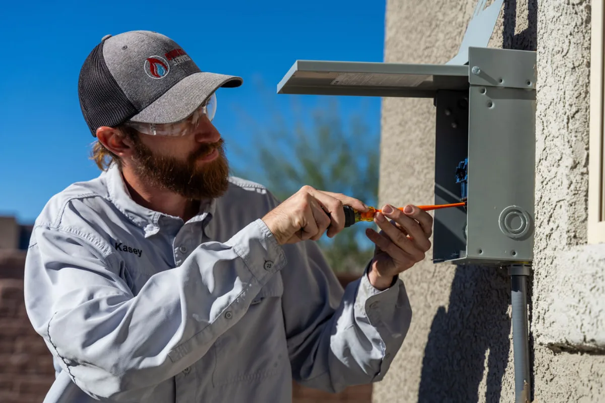 Technician in a Done Rite cap and safety glasses driving a screw inside a small grey outdoor disconnect on a stucco wall under blue sky