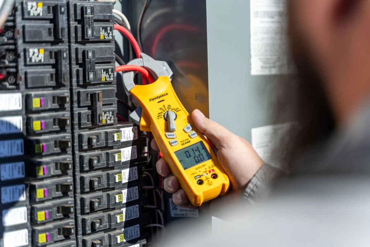 Yellow Fieldpiece clamp meter closed around a red service conductor inside a panel packed with black breakers