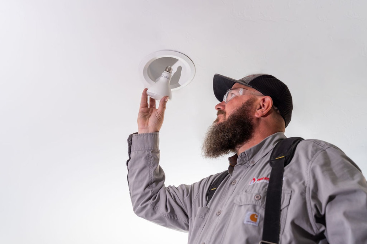 Bearded technician in a grey work shirt fitting an LED bulb into a recessed ceiling can light