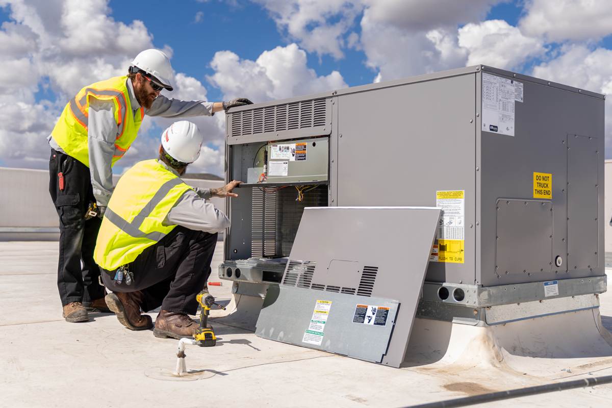 Two technicians in hard hats and hi-vis vests at a rooftop packaged unit with its coil compartment open, the access panel leaning on the roof and a cordless drill on the deck
