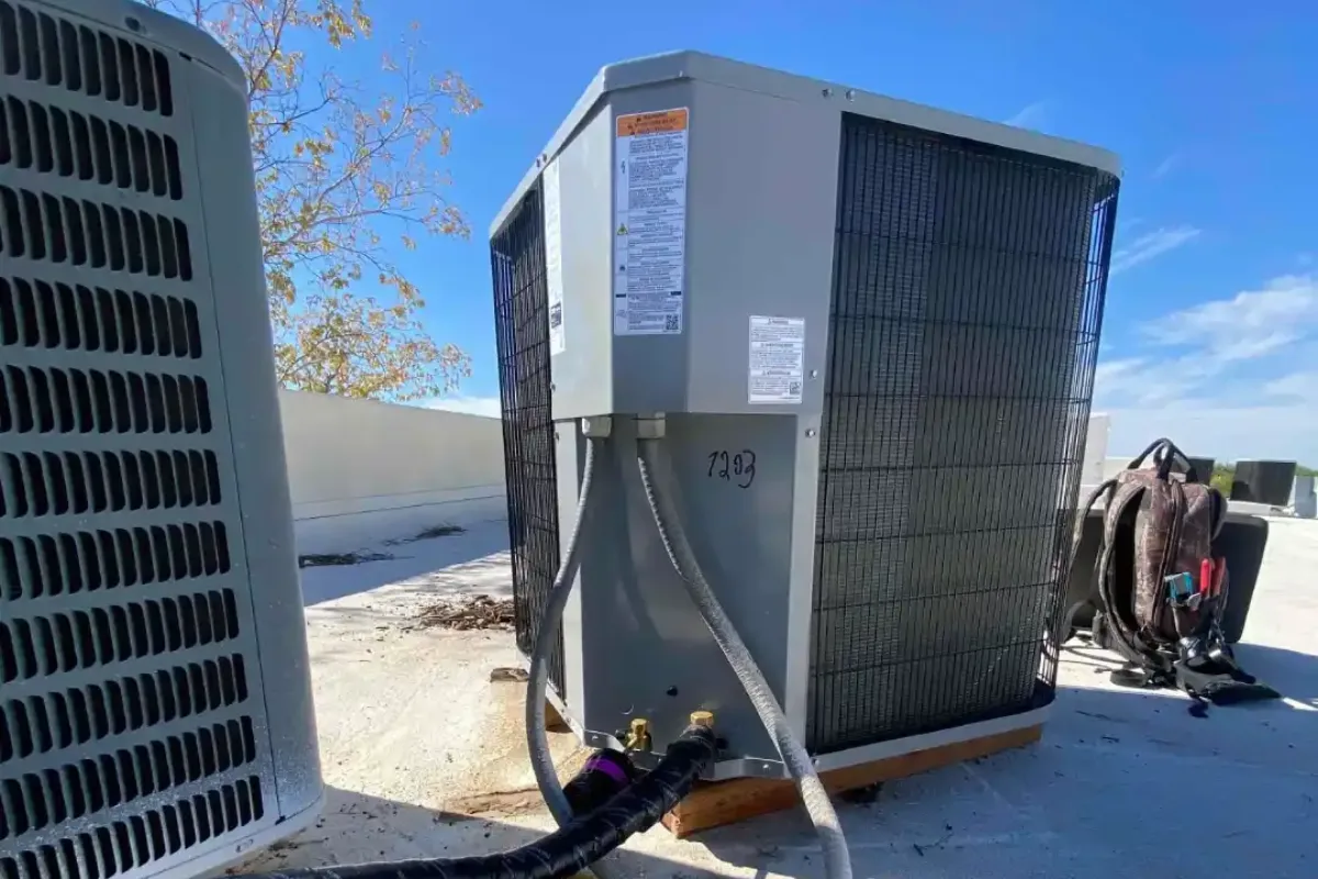 Outdoor condenser unit on a flat roof with insulated refrigerant lines running to the base and a tool bag set beside it