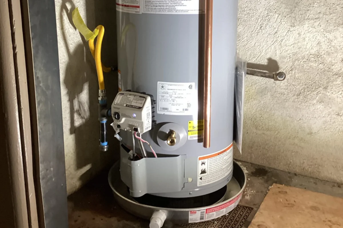 Grey gas water heater standing in a metal drip pan in a garage corner with a yellow flexible gas connector above