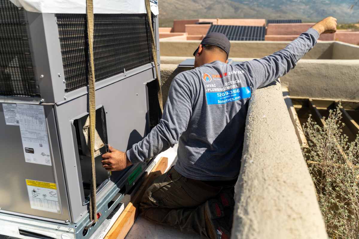 Technician in a branded Done Rite Services shirt kneeling beside a strapped condensing unit on a flat roof, tile-roofed houses and hillside beyond the parapet