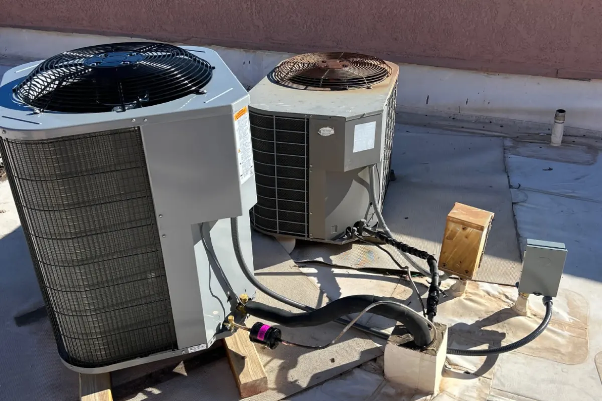 Two outdoor condenser units set on wood blocks on a flat roof with refrigerant lines and a disconnect box