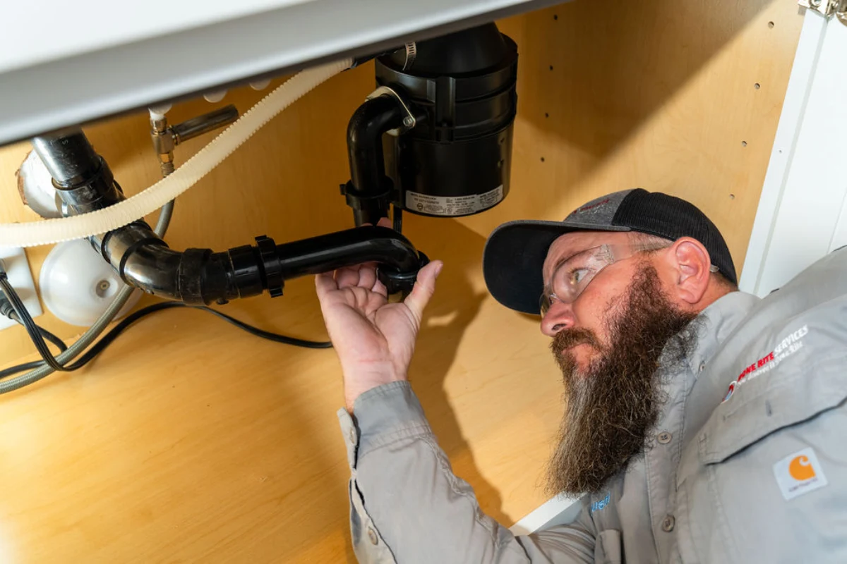 Bearded technician in a grey Done Rite Services shirt lying on his back inside a sink cabinet, cupping a hand under the black P-trap below a garbage disposal unit