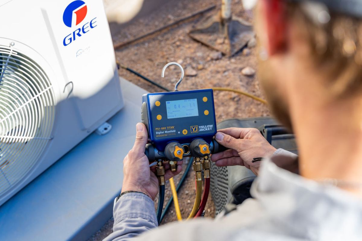 Technician holding a blue Yellow Jacket digital manifold gauge in front of a white Gree condenser on desert gravel