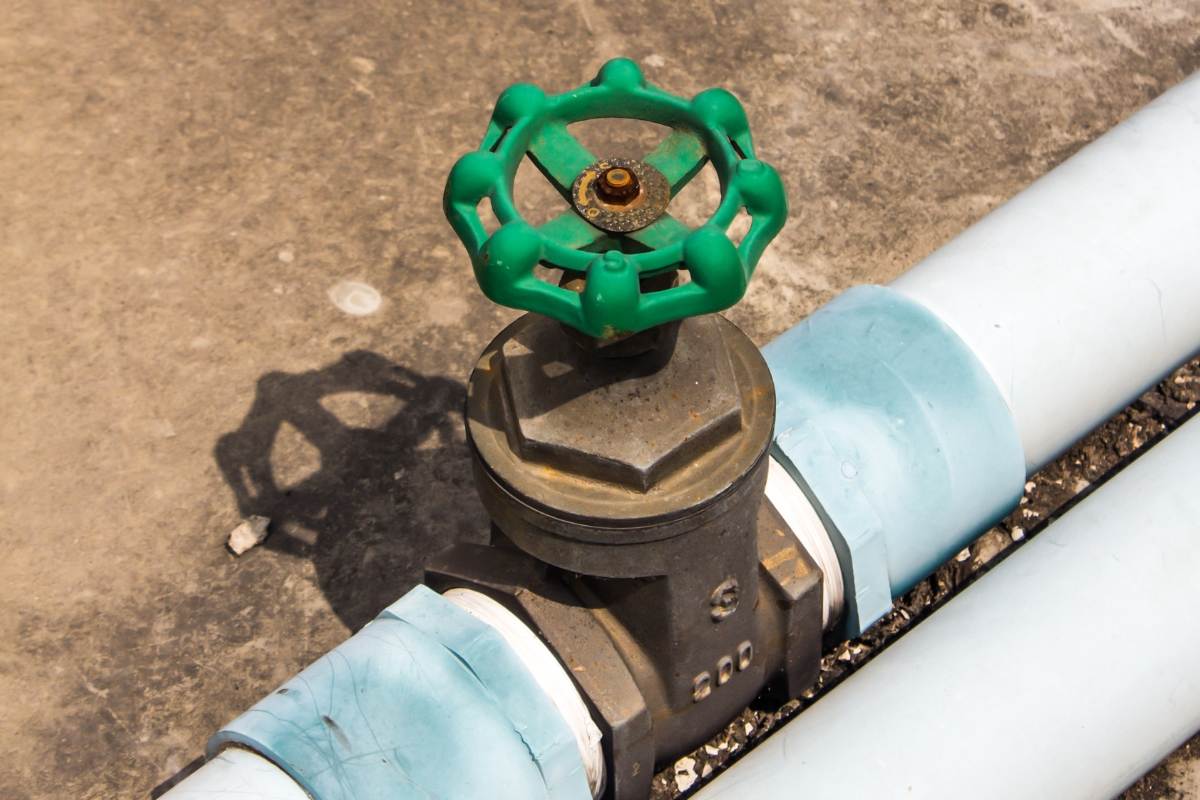 Green-handled gate valve joining two runs of white PVC pipe on bare ground