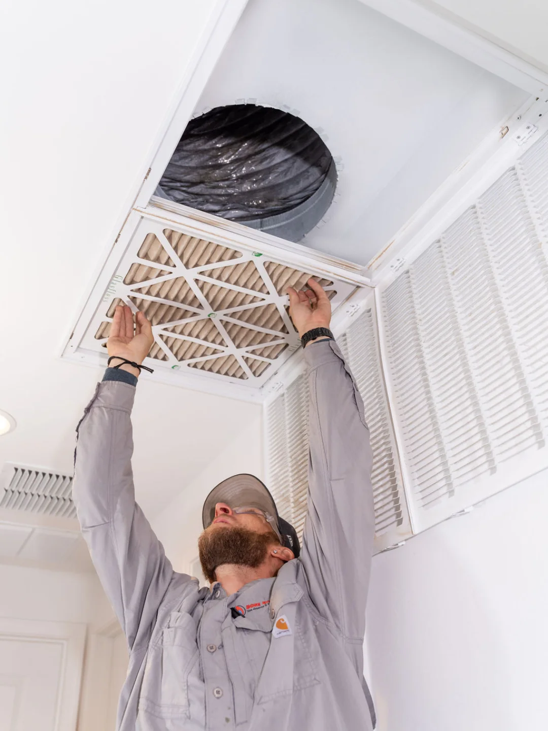 Technician in a grey Done Rite Services shirt reaching up to lift a pleated filter out of an open ceiling return grille, flexible duct visible in the cavity above