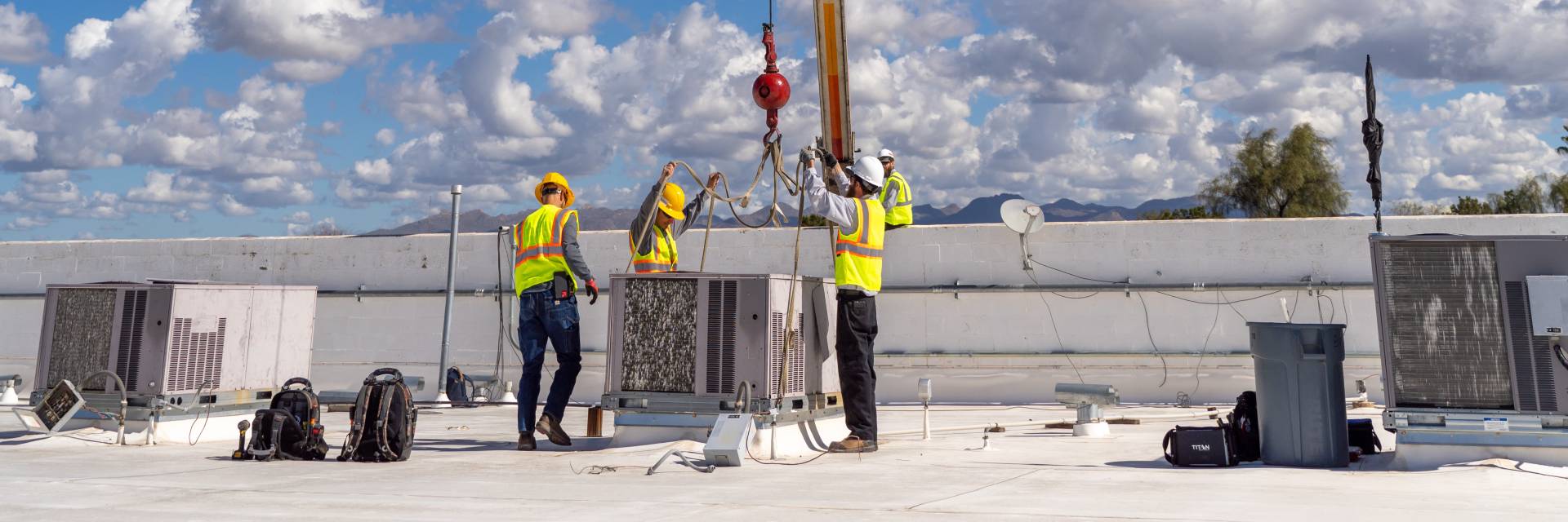 Crew in hard hats and hi-vis vests rigging a rooftop HVAC unit to a crane hook on a Tucson commercial building