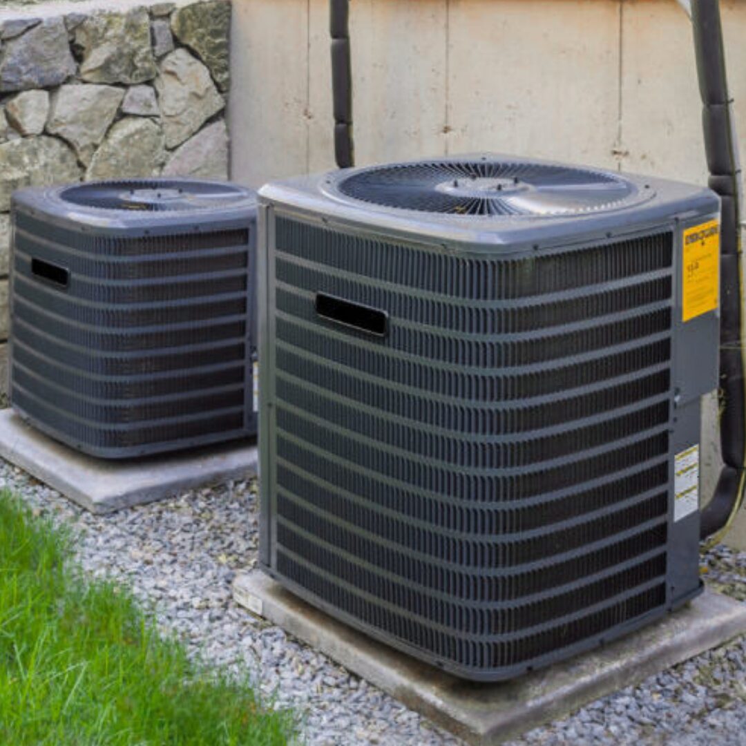 Two outdoor condensing units of different sizes sitting on concrete pads against a stucco wall beside a stone retaining wall, refrigerant lines running up behind them