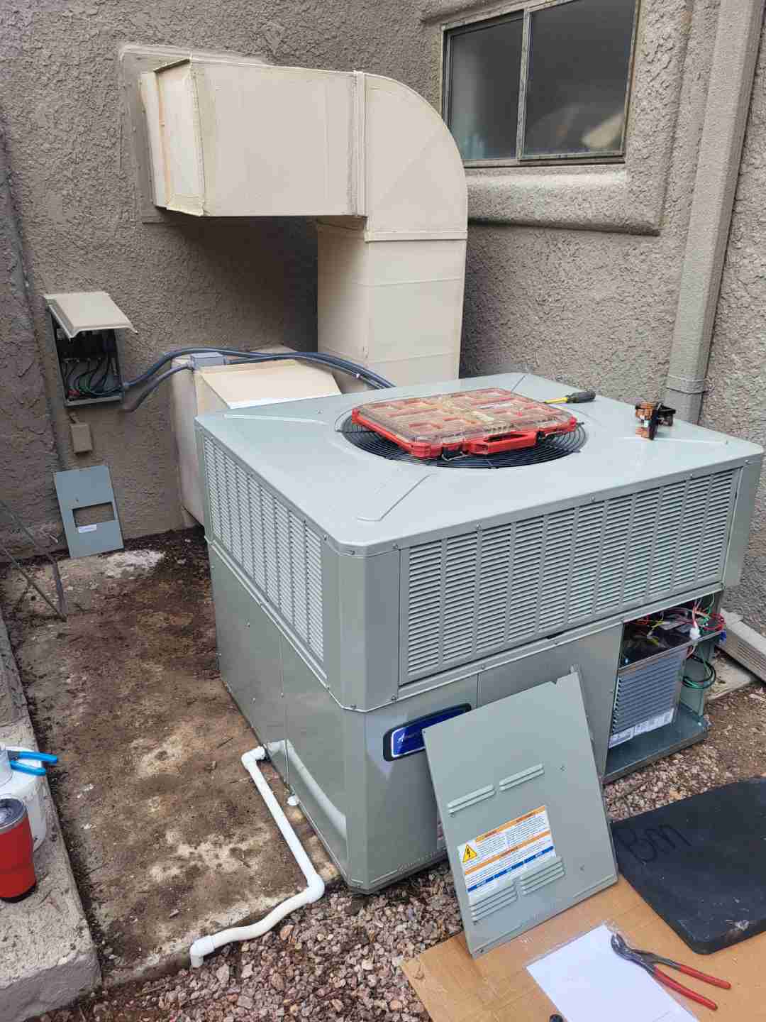 Residential packaged HVAC unit beside a stucco wall with its service panel removed and hand tools laid out on the ground