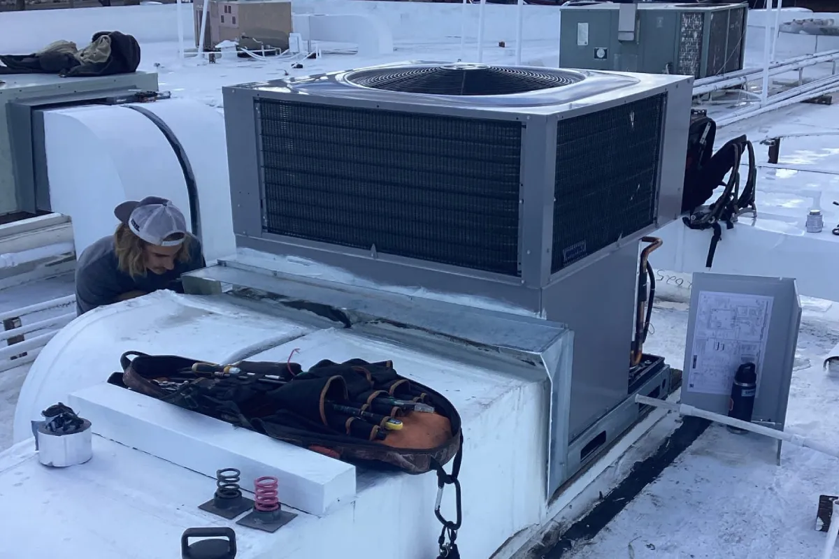 Technician working at the duct connection of a rooftop gas package unit, with copper gas piping running down to a disconnect box