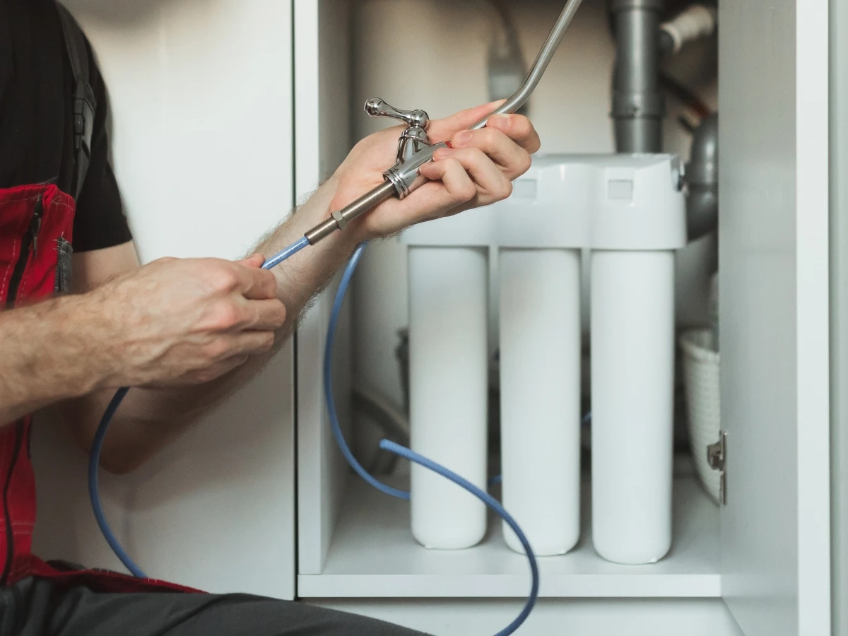 Technician connecting blue tubing and a chrome faucet stem to a three-stage under-sink filter housing inside a white cabinet