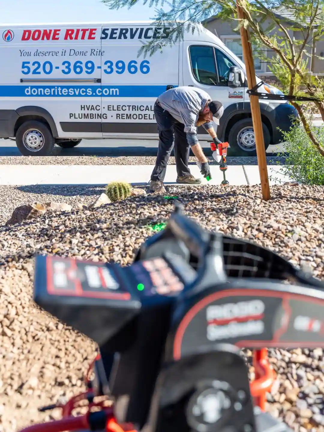 Technician bent over a gravel yard operating a locator wand with a Done Rite Services van parked at the kerb behind