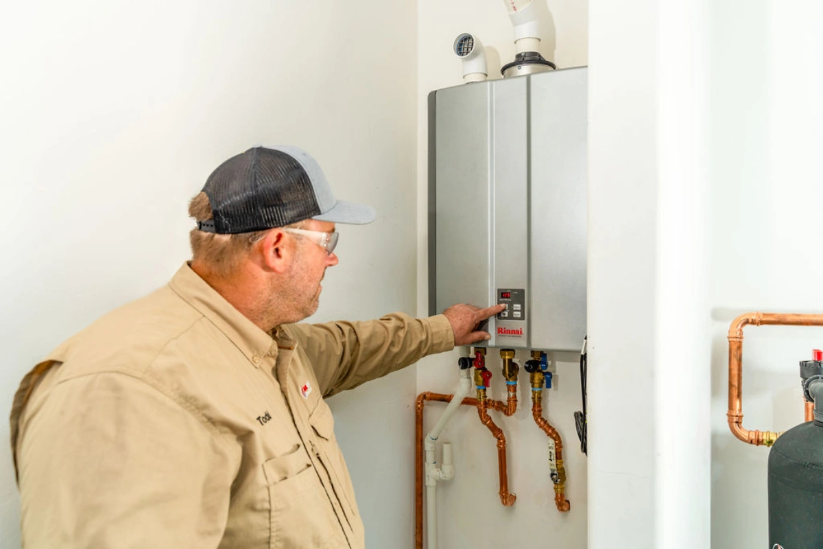 Technician in a tan work shirt pressing the control panel of a wall-mounted Rinnai tankless water heater with copper supply lines and isolation valves beneath it