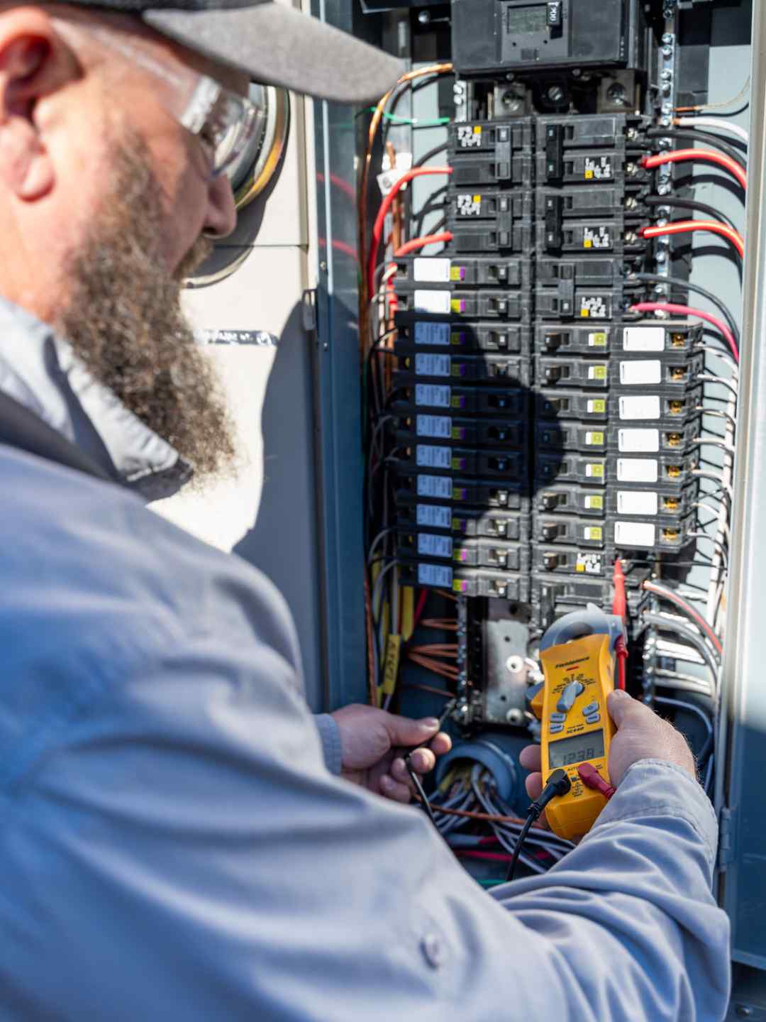 Electrician holding a yellow Fieldpiece meter reading 123.8 volts against the breakers of an open panel in bright sun