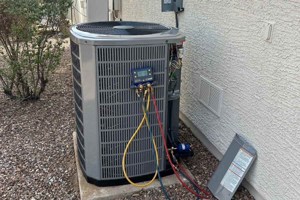Outdoor condensing unit on a concrete pad beside a stucco wall with refrigerant gauges and a blue digital manifold connected to the service ports