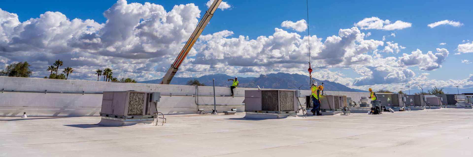 Crane boom lifting over a large white commercial roof with rows of package units and technicians in hi-vis working near the parapet